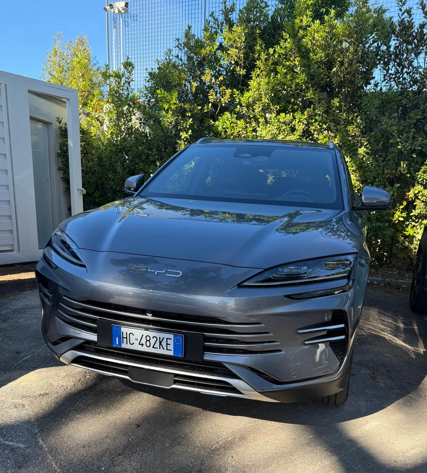 SUV BYD grigio della flotta DS Rentcar, targa HC 482KE, parcheggiato su asfalto davanti a una siepe verde