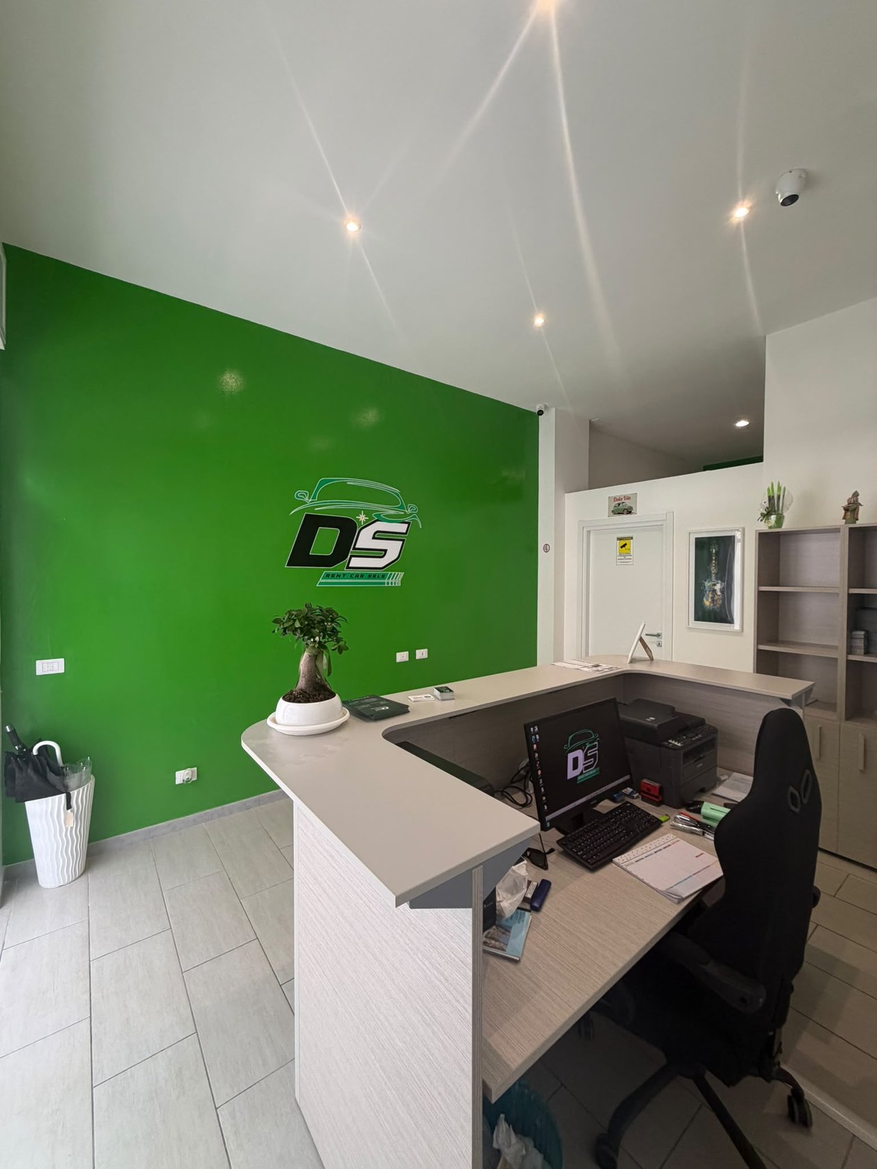 Reception di DS Rentcar con logo sulla parete verde, bancone grigio chiaro, monitor, bonsai e poltrona nera
